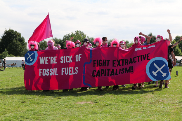 Eine Gruppe von Menschen auf einer grünen Wiese mit einer pinken Fahne, auf der steht: "Wir haben genug von fossilen Brennstoffen, Kampf gegen den extraktiven Kapitalismus", mit Zelten, Bäumen und einem klaren blauen Himmel im Hintergrund.