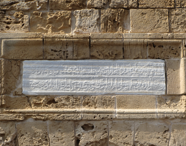 Ein Steintafel an einem Gebäude mit arabischer Schrift und kunstvollen Verzierungen, die wahrscheinlich eine Szene aus dem islamischen Glauben darstellen, umgeben von einem dekorativen Rahmen.