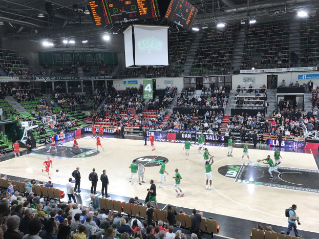Eine Gruppe von Menschen, die Basketball in einer vollen Halle spielen, mit Zuschauern auf den Tribünen und beleuchteter Spielfeldbeleuchtung.