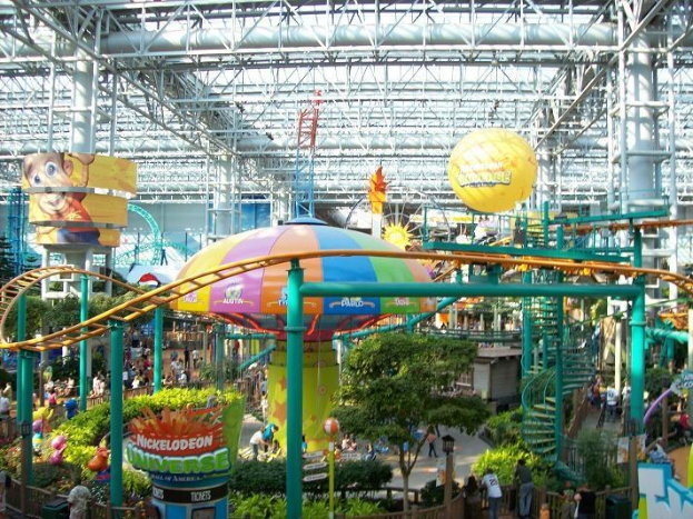 Ein großes Indoor-Wasserpark mit einer Achterbahn, umgeben von Menschen, Bäumen, Pflanzen und einer lebendigen Beleuchtung.