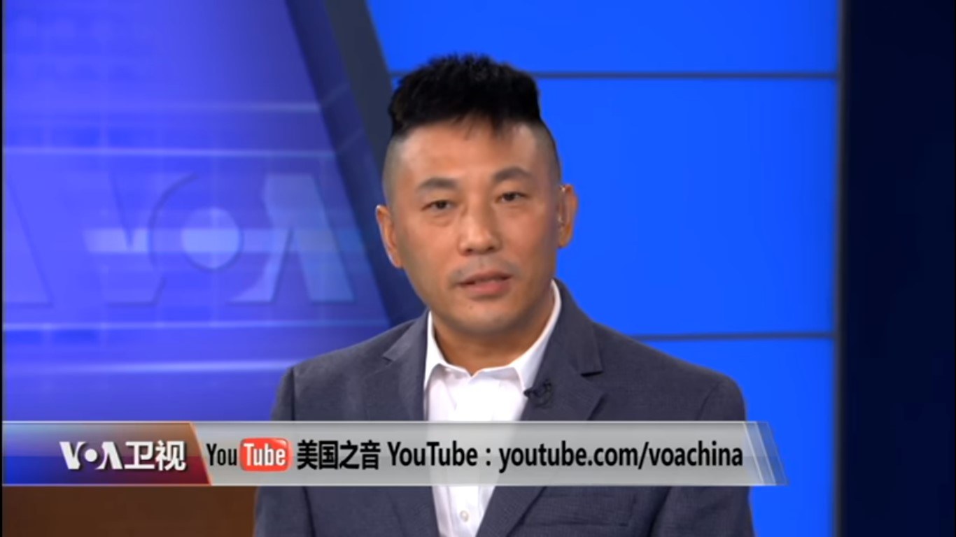 Ein chinesischer Nachrichtensprecher in Anzug und Krawatte erscheint auf einem Fernseher mit Text am unteren Rand und einem blauen Hintergrund.