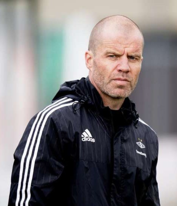 Mann mit Glatze in schwarzer Jacke, der wie ein Trainer aussieht, auf einem Fußballfeld stehend und eine marineblaue Jacke mit weißen Streifen und einem weißen Kragen tragend, die Hände verschränkt und einen ernsten Gesichtsausdruck.