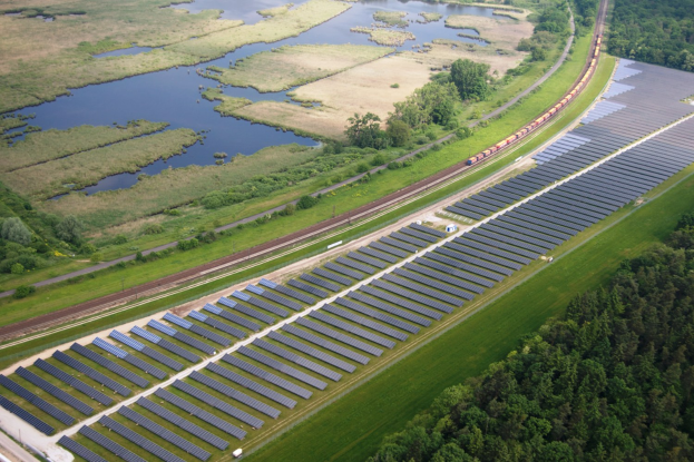 Luftaufnahme eines Solarparks mit Panelen, umgeben von Bäumen, Gras und Wasser, mit einem Zug auf einer nahen Schiene.