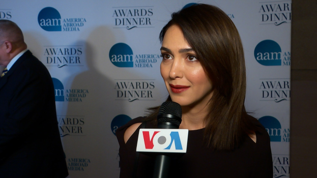 Emmy Rossum in einem schwarzen Kleid hält ein Mikrofon und steht neben einem Mann mit einem Banner 'Aam Awards Dinner' im Hintergrund.