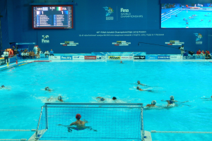 Eine Gruppe von Menschen spielt Wasserball in einem großen Pool mit einem Netz, Zuschauern auf der linken Seite und einem Bildschirm mit Text und Logos im Hintergrund bei den FINA-Weltmeisterschaften 2016 in Kazan.