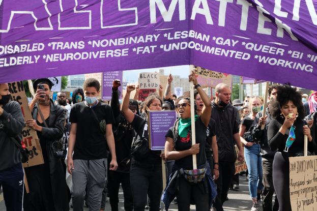 Eine Gruppe von Menschen marschiert mit Schildern und Transparenten die Straße entlang, einige tragen Masken und eine Person hält ein Mobiltelefon in der Hand, vor dem Hintergrund von Bäumen, Gebäuden und einem sichtbaren Himmel, wahrscheinlich bei einer Black-Lives-Matter-Demonstration in New York City.