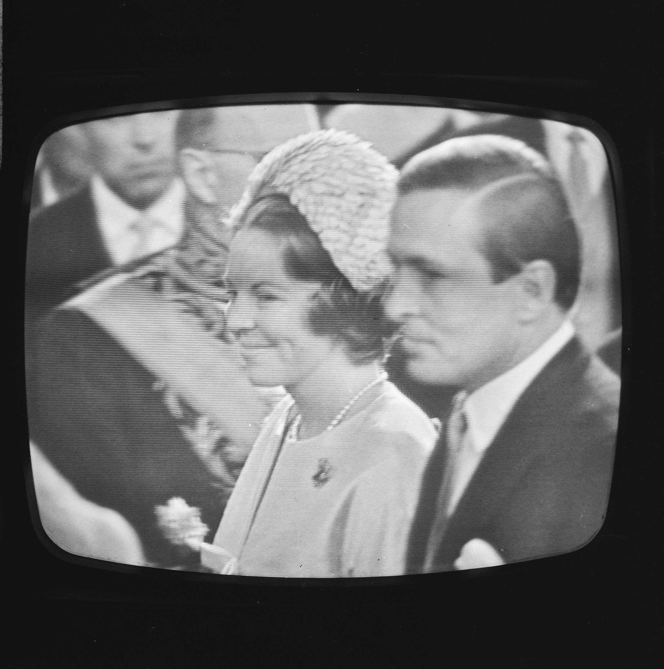 Schwarzer und weißer Fernseher, der einen Mann in Anzug und eine strahlende Frau zeigt, mit ein paar anderen Menschen im Hintergrund.