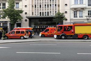 Gruppe von Feuerwehrautos auf einer Straße in Paris geparkt mit Menschen in der Nähe, Gebäuden, Bäumen und einem Fahrrad im Hintergrund, wahrscheinlich an einem Unfallort.