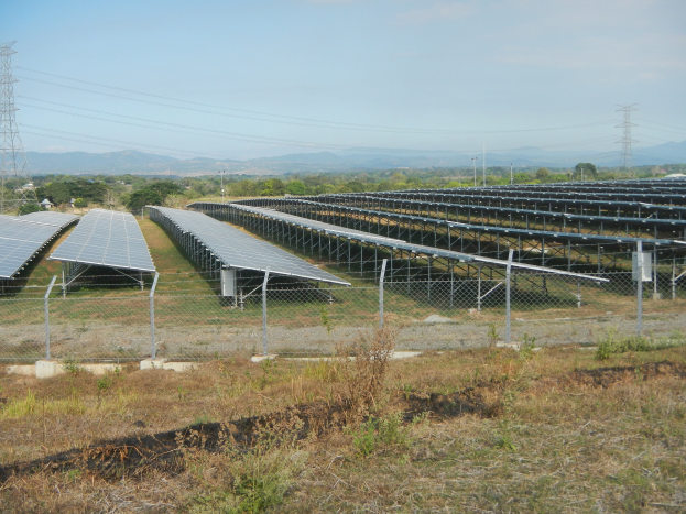 Luftaufnahme eines Solarpanelfelds, das von einem Zaun, Gras und Pflanzen umgeben ist, mit Bäumen, Übertragungsmasten, Stromleitungen und Hügeln unter einem klaren blauen Himmel.