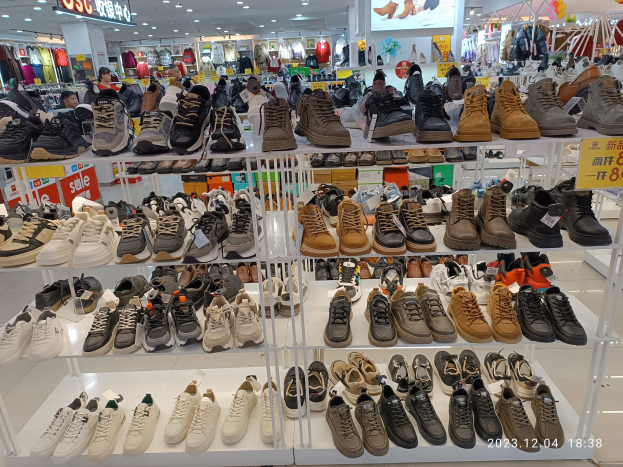Ein Schuhgeschäfts-Innenraum mit verschiedenen Schuhen auf Ständern, Preisschildern, Menschen im Hintergrund, hängenden Kleidern, Deckenlampen, einem Bildschirm und Texttafeln.