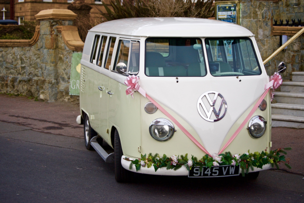 Ein weißer und rosafarbener VW-Bus mit floralen Verzierungen, der vor einer Steinmauer geparkt ist, neben einer Treppe und einem Gebäude im Hintergrund.