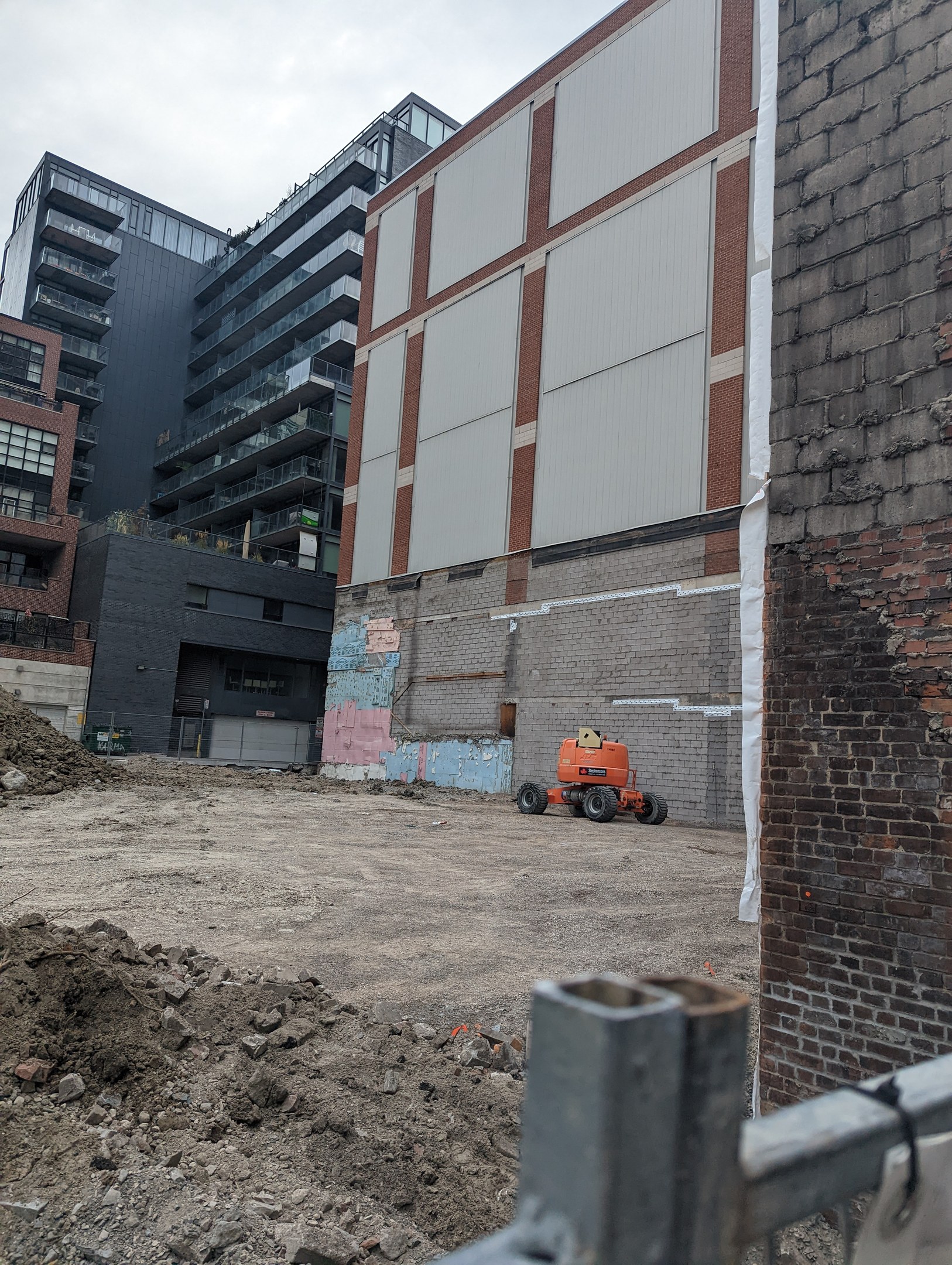 Baustelle mit einem teilweise abgerissenen Gebäude im Hintergrund, einem Fahrzeug auf dem Boden, einem Metallzaun und einem bewölkten Himmel, mit verstreuten Werkzeugen und Materialien, die auf Abrissaktivitäten hinweisen.