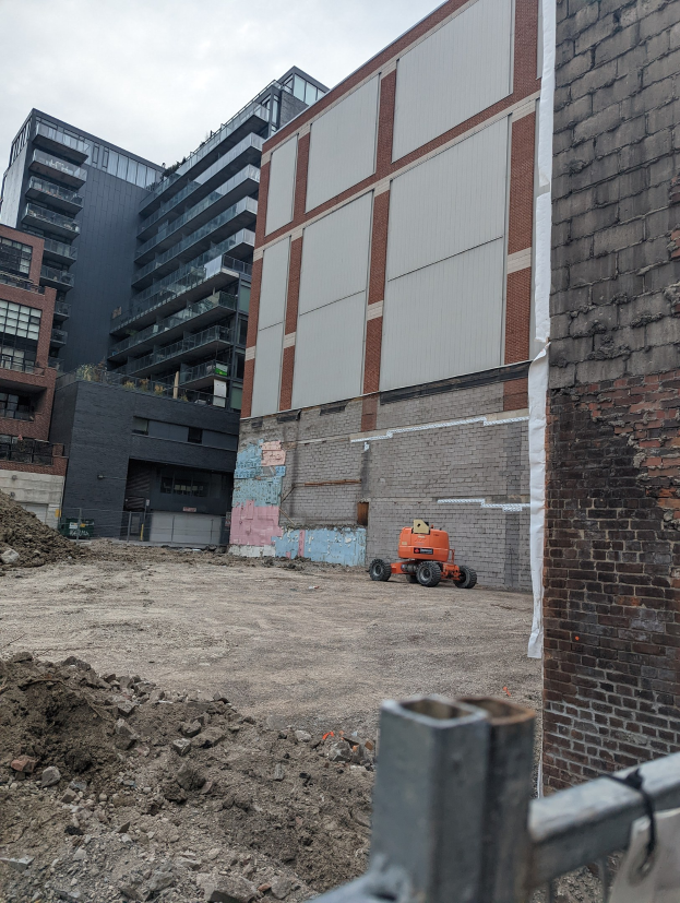 Baustelle mit einem teilweise abgerissenen Gebäude im Hintergrund, einem Fahrzeug auf dem Boden, einem Metallzaun und einem bewölkten Himmel, mit verstreuten Werkzeugen und Materialien, die auf Abrissaktivitäten hinweisen.