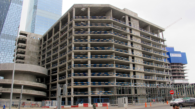 Ein großes Gebäude im Bau in einer städtischen Umgebung, umgeben von Wolkenkratzern, Fahrzeugen, Fußgängern, Verkehrsinfrastruktur und Kränen vor einem klaren blauen Himmel.