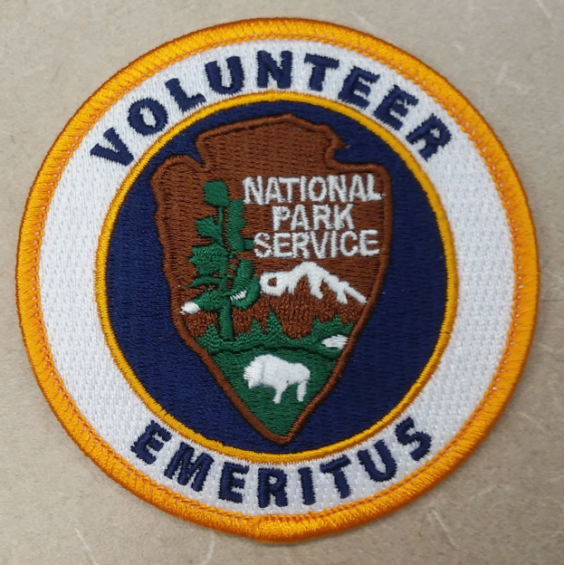 Ein rundes Abzeichen mit einem blauen Rand, einem weißen Hintergrund und fettem schwarzem Text, der "Volunteer National Park Service Emeritus" in der Mitte eines gelben Kreises lesen.