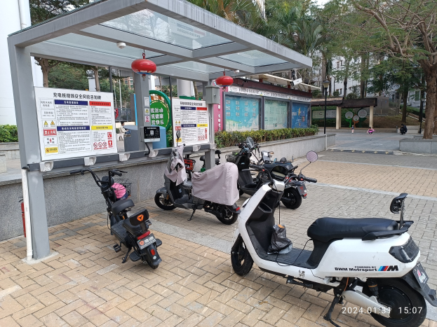 Elektrische Scooter vor einem Bushaltestellen mit nahen Informationsschildern, Bäumen, Gebäuden und einem klaren blauen Himmel.