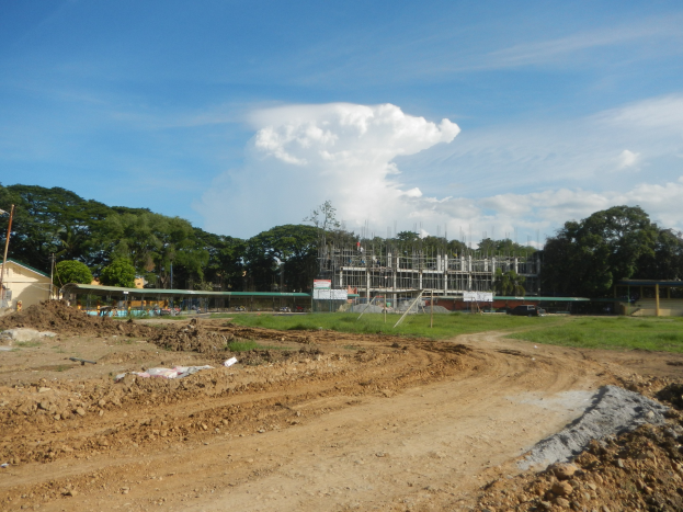 Ein Schotterweg führt zu einem teilweise errichteten Gebäude, das von Gras, Strommästen und Bäumen umgeben ist und unter einem bewölkten Himmel liegt.