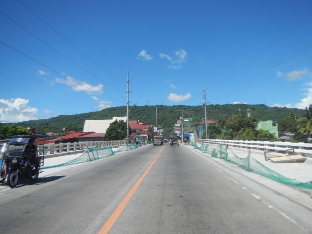 Autobahn mit fahrenden Fahrzeugen, ein stationäres Motorrad auf dem Seitenstreifen, Leitplanken, Strommasten mit Drähten, Bäume, ferne Gebäude, Hügel und ein bewölkter Himmel.