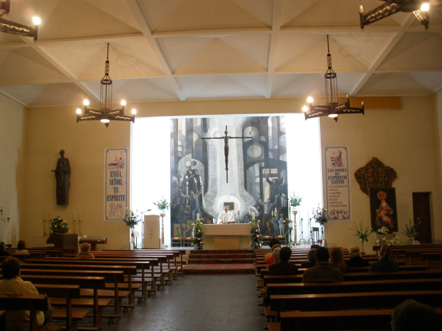 Eine Kircheninnenseite mit Menschen in Kirchenbänken, einer Person an einem Podium mit Mikrofon, Kronleuchtern, Blumenbouquets, Kerzen und Postern an den Wänden.