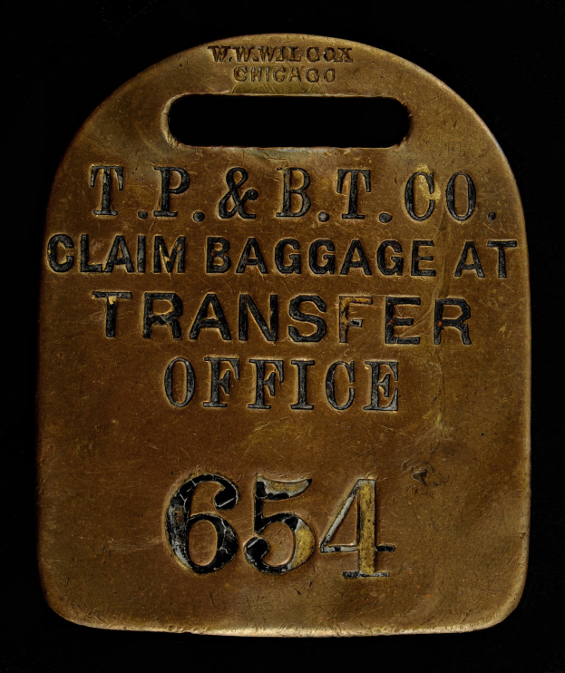 Brass luggage tag with "T.P. & B.T. Co. Claim Baggage at Transfer Office" engraved on it against a dark background.