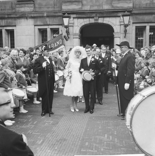 Ein Brautpaar geht eine Straße entlang vor einer Menge, einige halten Musikinstrumente, mit einem Gebäude und einem Banner im Hintergrund, alles in Schwarzweiß dargestellt.
