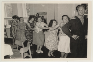 Eine Gruppe von Menschen, die in einem Raum im Stil der 1950er Jahre tanzen, mit Stühlen, einem Tisch, einem Foto in einem Rahmen an der Wand und Fenstern im Hintergrund.