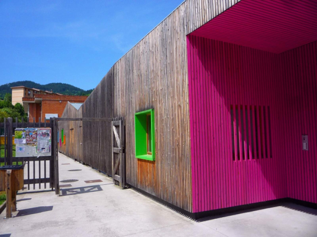 Rosa Gebäude mit grüner Tür und Fenstern, hölzerner Zaun, Pappschild, Mülltonne, Gras, Bäume, entfernte Gebäude, Hügel und bewölkter Himmel, Teil eines neu umgebauten Schulspielplatzes in Spanien.