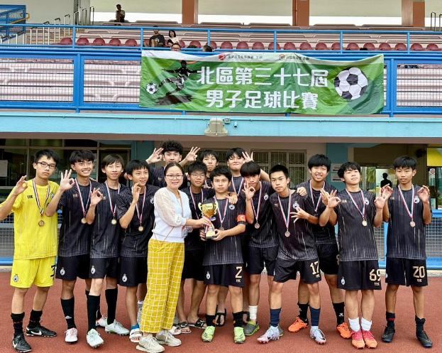 Gruppe junger Männer in Fußballtrikots auf einem Feld stehend, Medaillen tragend und einen Pokal haltend, mit einem "Yokohama U-16 Jungenfußballteam"-Schild im Hintergrund und sitzenden Zuschauern.