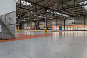 Großes Industriegebäude mit zahlreichen Fenstern, Metallstützen, einer Treppe mit Geländer, einem Schild, Deckenleuchten und zusätzlichen Stützen.