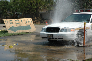Polizeiauto fährt durch eine Pfütze mit verstreuten Gegenständen, darunter eine Tafel mit Text und ein Verkehrskegel, Bäume, Gebäude und einen klaren blauen Himmel im Hintergrund.
