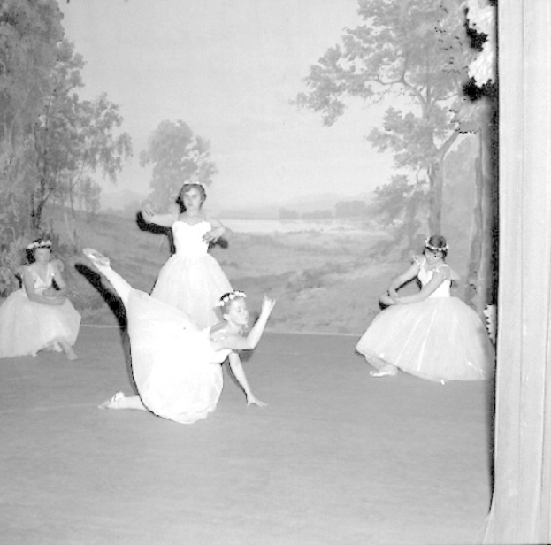 Vier junge Balletttänzerinnen in weißen Kleidern tanzen auf einer Bühne mit einer gemalten Bäume-und-Himmel-Bühne, in Schwarz-Weiß.