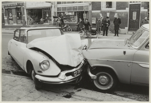 Schwarz-weißes Foto von zwei beschädigten Autos am Straßenrand mit Menschen und Fahrrädern in der Nähe, Gebäude im Hintergrund.