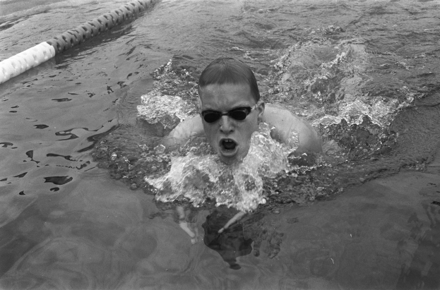 Ein Mann schwimmt in einem Pool und trägt eine Schwimmbrille, mit offenem Mund, umgeben von Bahnmarkierungen, auf einem Schwarz-Weiß-Bild.