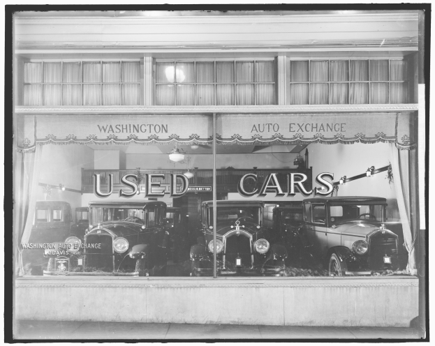Schwarz-weiß-Foto eines Gebrauchtwagenhändlers in Washington, D.C., mit durch die Glasscheibe sichtbaren Autos und beleuchteter Wandtext.