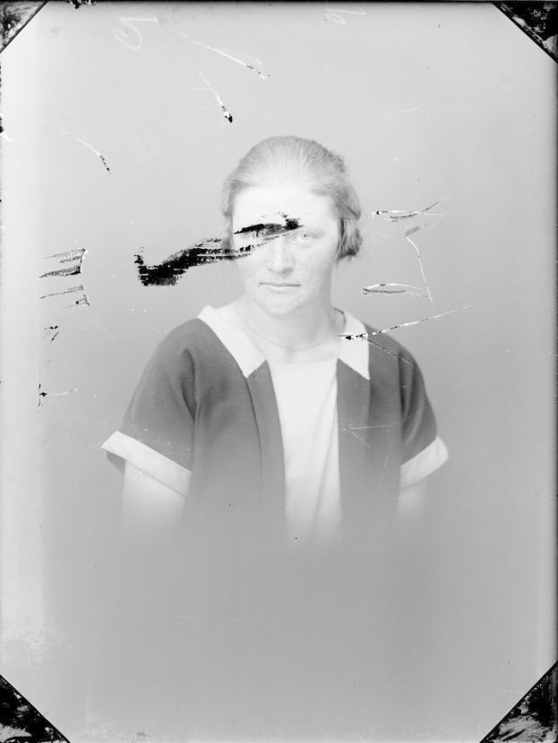 Schwarz-weiß-Foto einer Frau mit einem zerbrochenen Glasfenster im Hintergrund, ihr Gesicht ist deutlich zu sehen, mit direktem Augenkontakt und leicht geöffneten Lippen, das Haar zurückgekämmt, die Hände vor der Brust verschränkt.