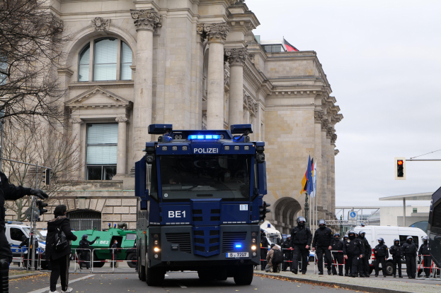 Gruppe von Polizeibeamten vor einem großen Gebäude mit Fenstern, Säulen und Bögen, mit Fahrzeugen auf der Straße und einer Person mit einer Kamera links.