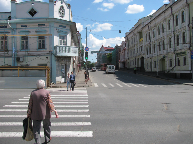 Alte Frau überquert die Straße vor einem Gebäude, mit Fußgängern, Fahrzeugen, Schildern, Strommasten, Bäumen und einem bewölkten Himmel im Hintergrund.