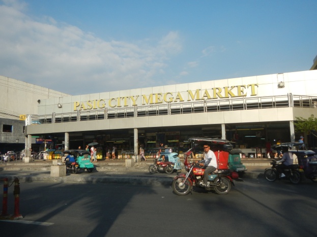 Eine belebte Straßenszene am Pasig City Mega Market mit Fahrzeugen und Fußgängern, ein Gebäude mit sichtbarem Text im Hintergrund und ein bewölkter Himmel.