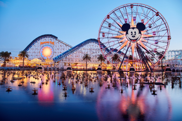 Eine lebendige Dämmerungsszene im Disneyland California Adventure mit einem riesigen Mickey-Maus-Rad im Vordergrund, einer Achterbahn im Hintergrund und Booten auf dem Wasser, mit einem Himmel aus Orangen, Pink- und Purton und silhouettenartigen Bäumen.