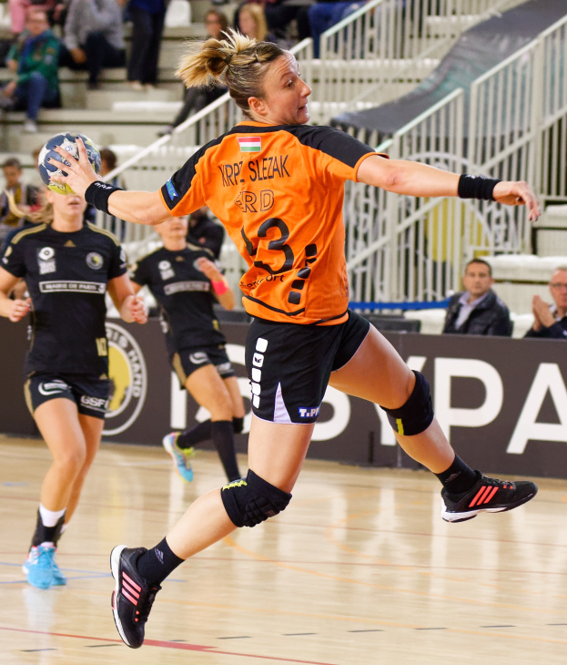 Eine Frau in einem orangen T-Shirt und schwarzen Shorts hält einen Handball, mit Zuschauern auf Treppen und Geländern im Hintergrund während der "Krp Slezak - Handball-Weltmeisterschaft 2019".