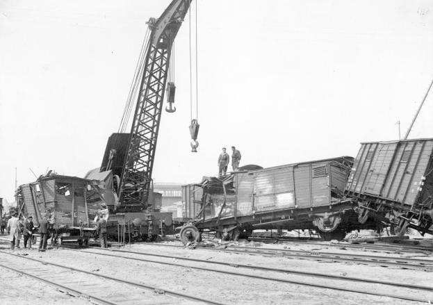 Schwarzes und weißes Bild einer Zugkatastrophe mit einem Kran, der einen Zugwagen auf die Schienen hebt, mehrere Menschen in der Nähe, Gebäude und ein klarer Himmel im Hintergrund.