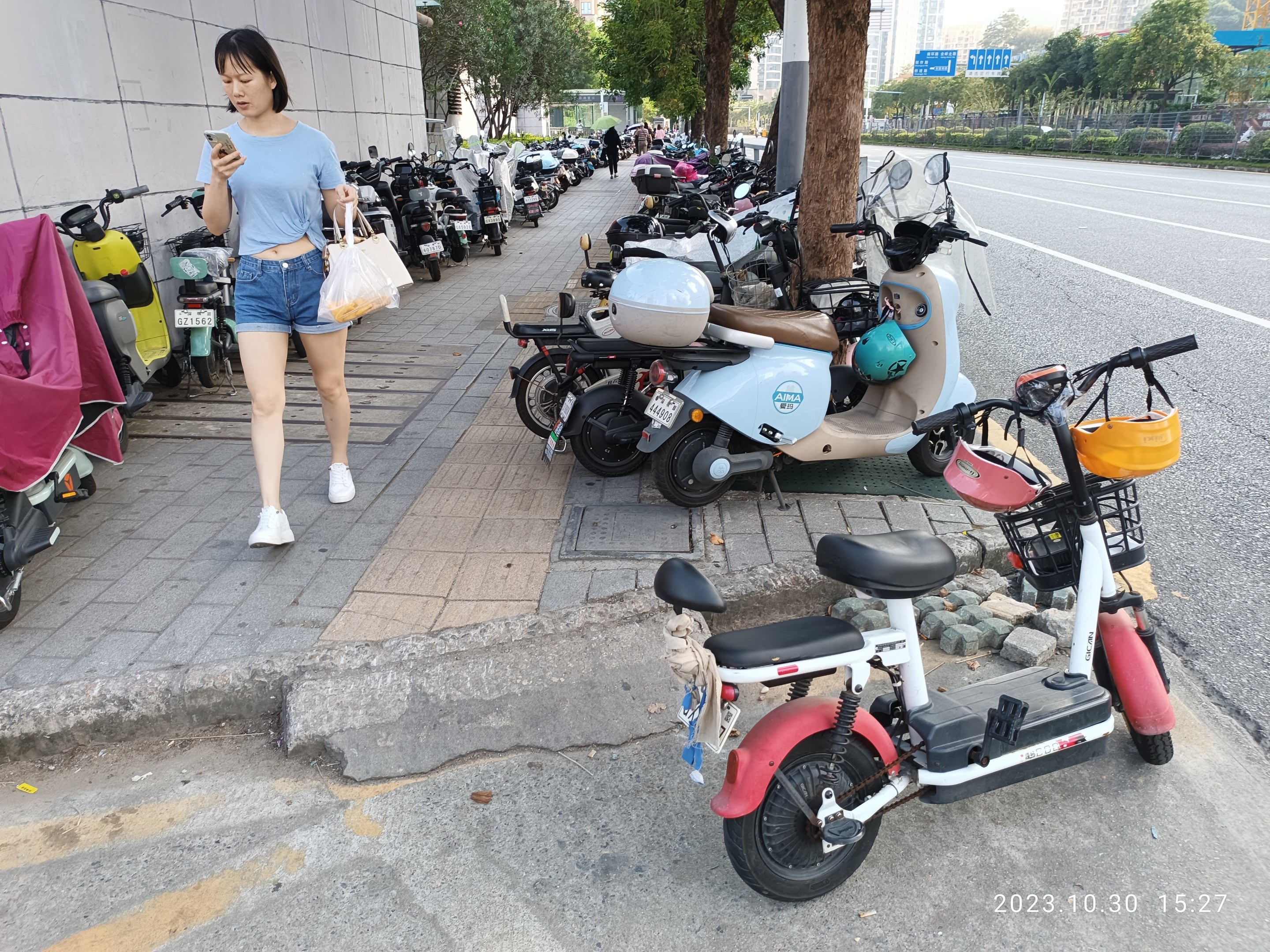 Eine Frau in einem blauen T-Shirt, blauen Shorts und weißen Schuhen geht auf einem Bürgersteig, hält ein Handy und eine Tasche und läuft an einer Reihe geparkter E-Scooter vorbei, mit Bäumen, Gebäuden und einem klaren blauen Himmel im Hintergrund.
