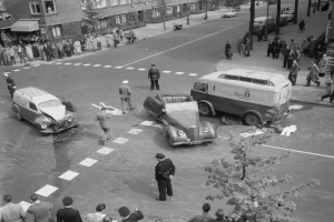 Schwarz-weiß-Szene eines Autounfalls an einer Straßenkreuzung mit mehreren Fahrzeugen, darunter ein Lieferwagen, und einer Gruppe von Menschen in der Nähe, mit Gebäuden, Bäumen, Pfählen und Schildern im Hintergrund.