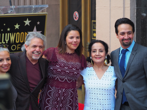 Gruppe von Menschen auf einem roten Teppich bei der Zeremonie des Hollywood Walk of Fame posierend, mit einer Kamera auf der linken Seite und einer Tafel, einem Podium und einer Wand im Hintergrund.