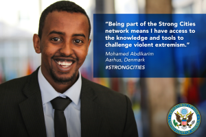 Ein Mann im Anzug und Krawatte lächelt in die Kamera, mit einem Logo und Text auf der rechten Seite, der besagt "Teil des starken Städtenetzwerks zu sein bedeutet, Zugang zu Wissen und Werkzeugen zu haben, um gewalttätigen Extremismus herauszufordern", und einem unscharfen Hintergrund.