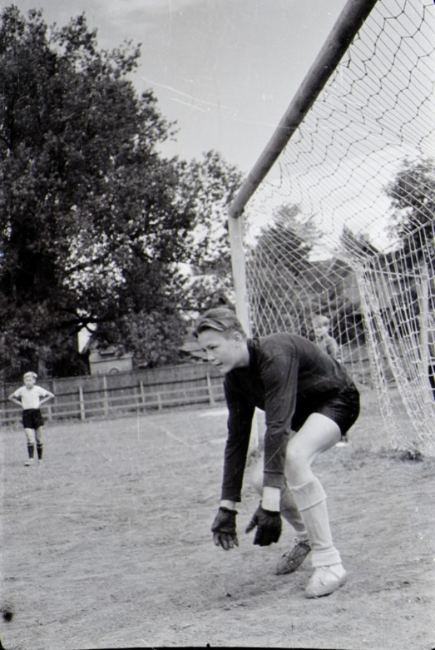 Ein Schwarz-Weiß-Foto eines jungen Jungen, der auf einem Feld Fußball spielt, mit einem Zaun, Bäumen und einem klaren Himmel im Hintergrund, anderen Kindern, die in der Ferne spielen, und einer Personenschuhwerk im Vordergrund.