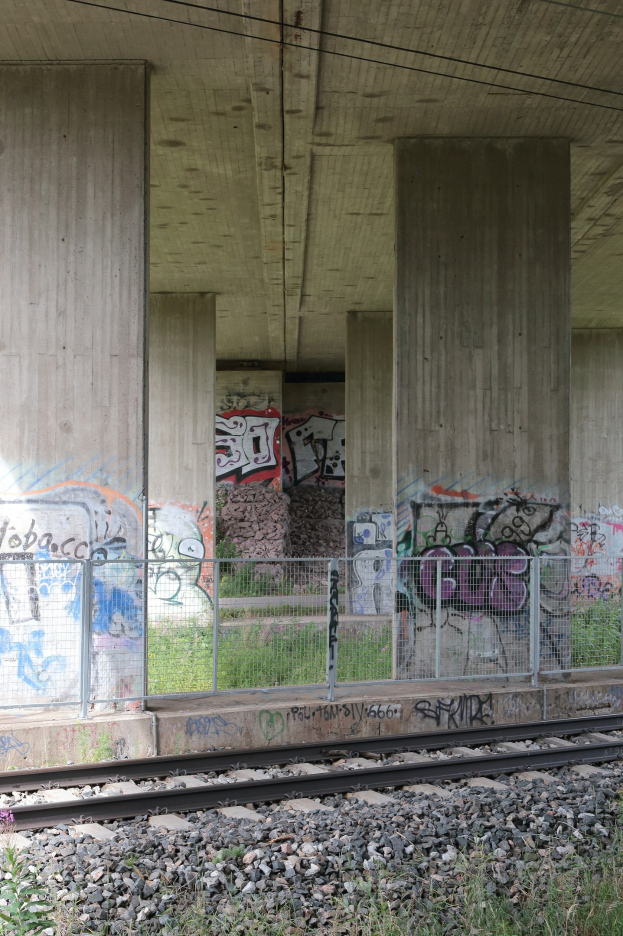 Eisenbahnschiene mit Graffiti an der Seite, umgeben von Steinen, Gras, einem Zaun, Säulen, Drähten und einem Dach.