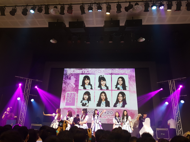 Eine Gruppe von Menschen, die auf einer hell erleuchteten Bühne performen, mit einer Menge darunter, einschließlich eines Sängers mit einem Mikrofon und eines Musikers, der ein Instrument spielt, während ein großer Bildschirm Bilder von BK48 anzeigt.