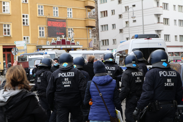 Polizisten in Uniform vor einer Menge während einer Demonstration in Berlin, mit Fahrzeugen, Gebäuden und einem Banner im Hintergrund.