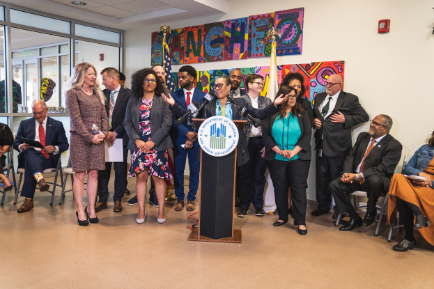 Gruppe von Menschen vor einem Rednerpult mit einem Mikrofon und einer Tafel mit der Aufschrift "Sancho-Stadtrat tritt dem Stadtrat bei", mit sitzenden und stehenden Teilnehmern, Fahnen, einem Feuerlöscher und Deckenleuchten.